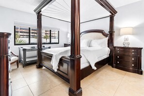 A four-poster bed with a dark finish, a nightstand with a lamp, and a view of the outside through a window.