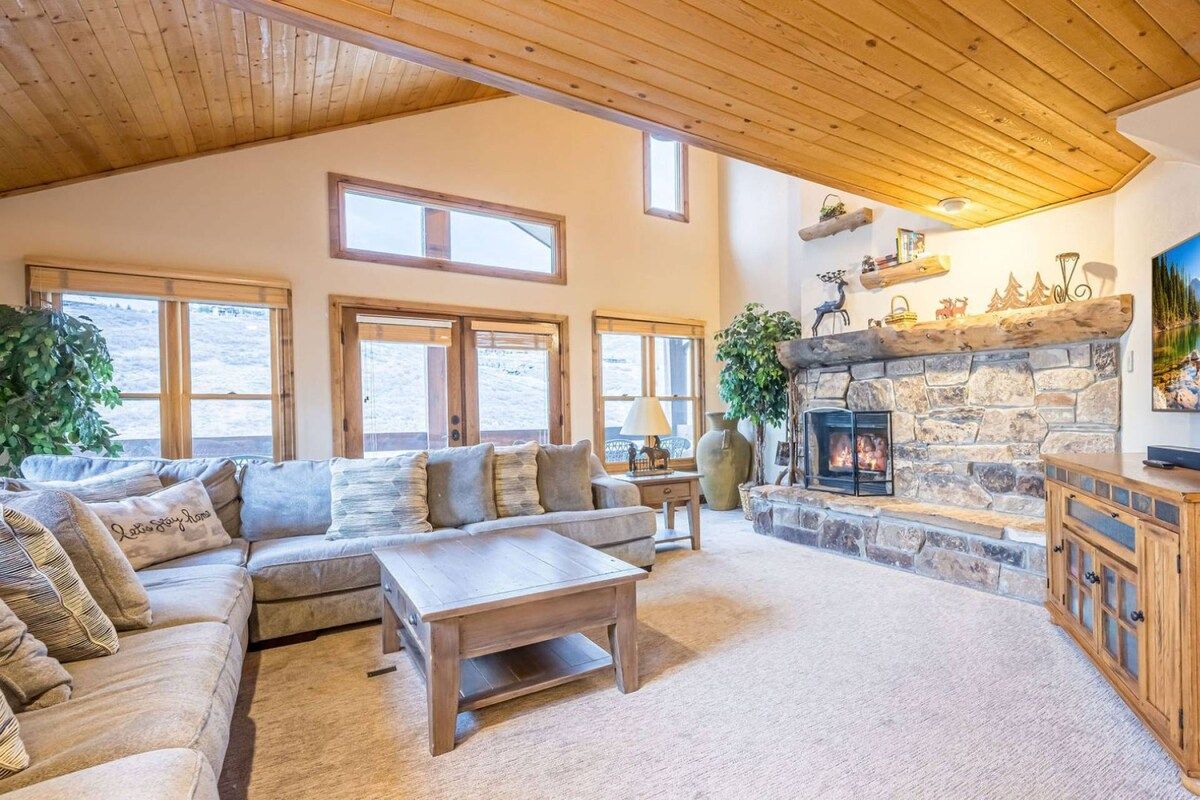 A cozy living room with a stone fireplace, large windows, and a high ceiling.