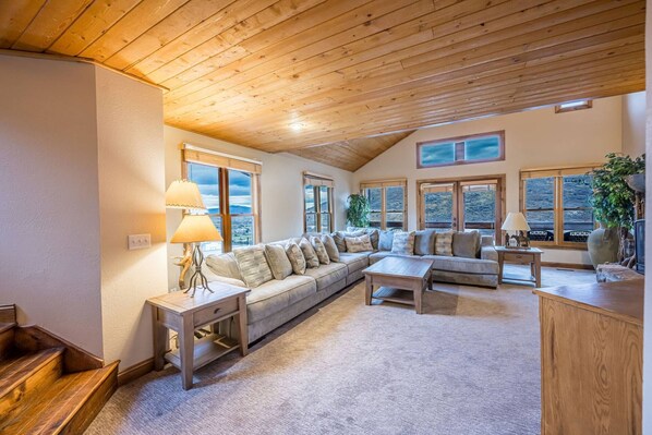 A spacious living room with a wooden ceiling, large windows offering a view of the outdoors, a sofa set, a coffee table, and a lamp.