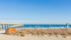 Ein Strand mit einer hölzernen Seebrücke, die ins Meer führt, ein sandiger Vordergrund mit trockenem Gras und ein klarer, blauer Himmel.