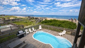 Una terraza de madera con piscina, sillas de descanso blancas y vista a la playa y al océano.