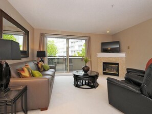Ein modernes Wohnzimmer mit Sofa, Sessel, Fernseher, Kamin und einem Balkon mit Blick auf Gebäude.