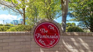 Um letreiro vermelho com o texto 'The Peninsula', rodeado por um muro de tijolos e árvores.
