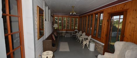 Une véranda couverte aux murs en bois et aux grandes fenêtres, aménagée avec des chaises blanches, un canapé et une petite table.