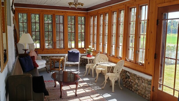 Une pièce baignée de lumière, avec des murs en bois, de grandes fenêtres, une cheminée en pierre et un coin salon confortable, comprenant une table et des chaises.