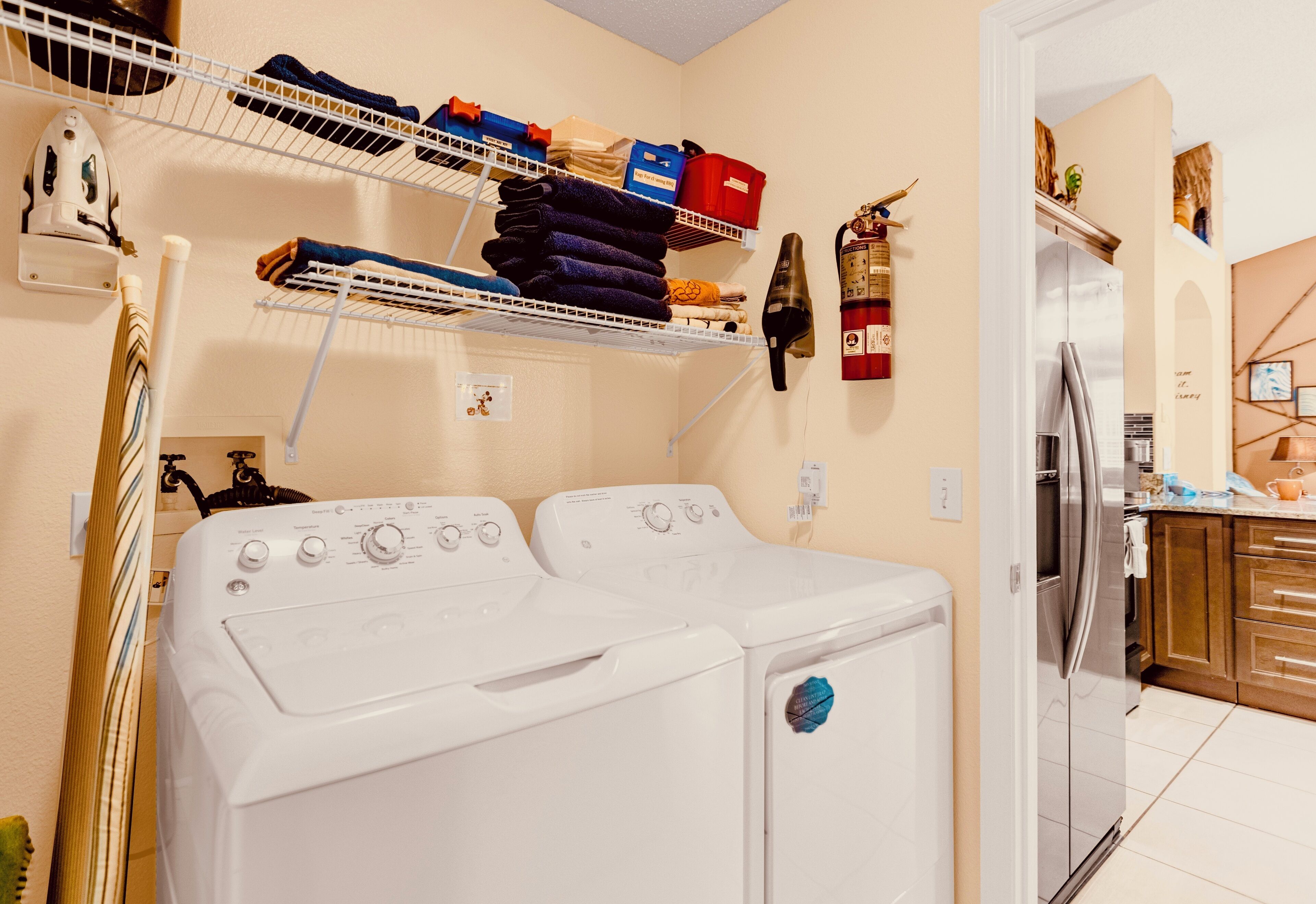 A laundry area with two white front-loading washers, a shelf with folded towels and storage boxes, a fire extinguisher, and a kitchen area visible through an open doorway.
