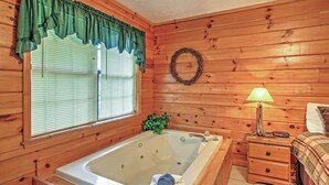 Interior de una cabaña de madera con una bañera grande, una ventana con persianas, una mesita de noche con una lámpara y una cama con manta escocesa.