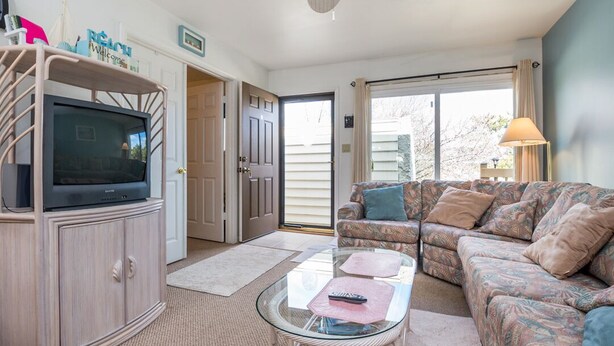 A living room with a TV on a cabinet, a glass coffee table, a patterned sofa, and a ceiling fan.