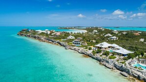 An aerial view of a coastal resort with clear turquoise waters, a rocky shoreline, and several buildings with swimming pools. - Villa Calypso: Ocean Access, Pool, Kayaks, & More! (Providenciales)