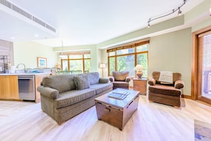 A spacious living area with a sofa, armchairs, and a coffee table. A kitchen with a dishwasher and cabinets is visible in the background.