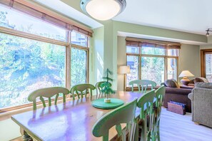 A dining area with a wooden table and chairs, a small Christmas tree, and large windows with a view of trees.