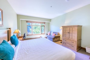 A bedroom with a large bed, a wooden wardrobe, a window with a view of greenery, and a desk with a lamp.