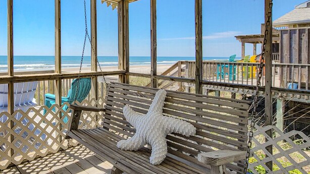 Une terrasse en bois avec un banc et une décoration en forme d’étoile de mer, donnant sur une plage sous un ciel dégagé.