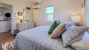 Une chambre à coucher comprenant un lit, des oreillers, une table de chevet, une lampe, un ventilateur de plafond, un appareil de climatisation, une commode, un miroir, une fenêtre et une porte donnant sur une salle de bain.