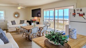 Un salon chaleureux avec une table à manger, des chaises en osier, un canapé blanc et une grande fenêtre donnant sur la plage.
