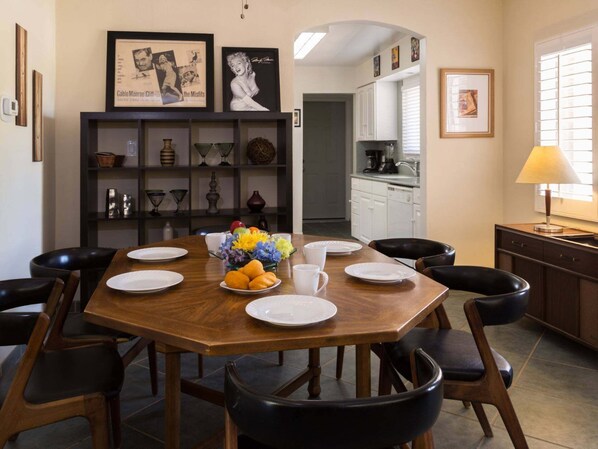Un coin repas avec une table en bois, des chaises noires et un vase de fleurs. Des tableaux encadrés ornent le mur, et on aperçoit une cuisine au fond.