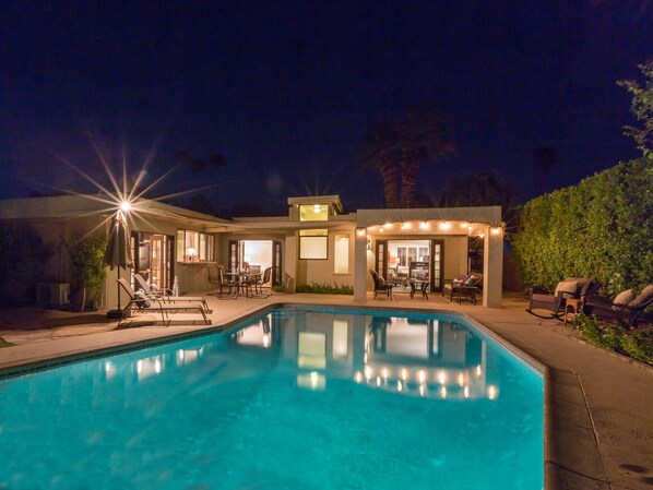 A swimming pool with a sunken seating area, a covered patio with outdoor furniture, and a well-lit entrance. - Reeves Garden House – Celebrity Pedigree (Palm Springs)