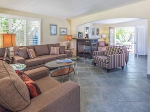 A living room with a sofa, armchair, and coffee table. There is a fireplace and a door leading to an outdoor area. - Reeves Garden House – Celebrity Pedigree (Palm Springs)