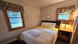 A bedroom with a bed, a nightstand, a lamp, a bench, and two windows with curtains. - Family resort cottage 3 on Big Glen Lake, Rent 1 or 5, dock, kayaks, rowboat (Maple City)
