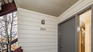 Un porche de madera con el número "1823" en la pared, una puerta oscura y un felpudo con diseño geométrico.