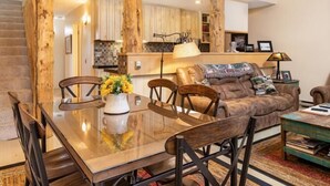 Amplia sala de estar con una mesa de comedor de madera, sillas y un sofá. Un candelabro cuelga del techo, y una escalera conduce a un nivel superior.