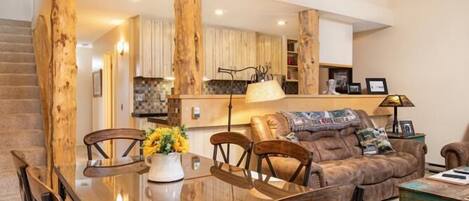 A spacious living area with a wooden dining table, chairs, and a sofa. A chandelier hangs from the ceiling, and there is a staircase leading to an upper level.