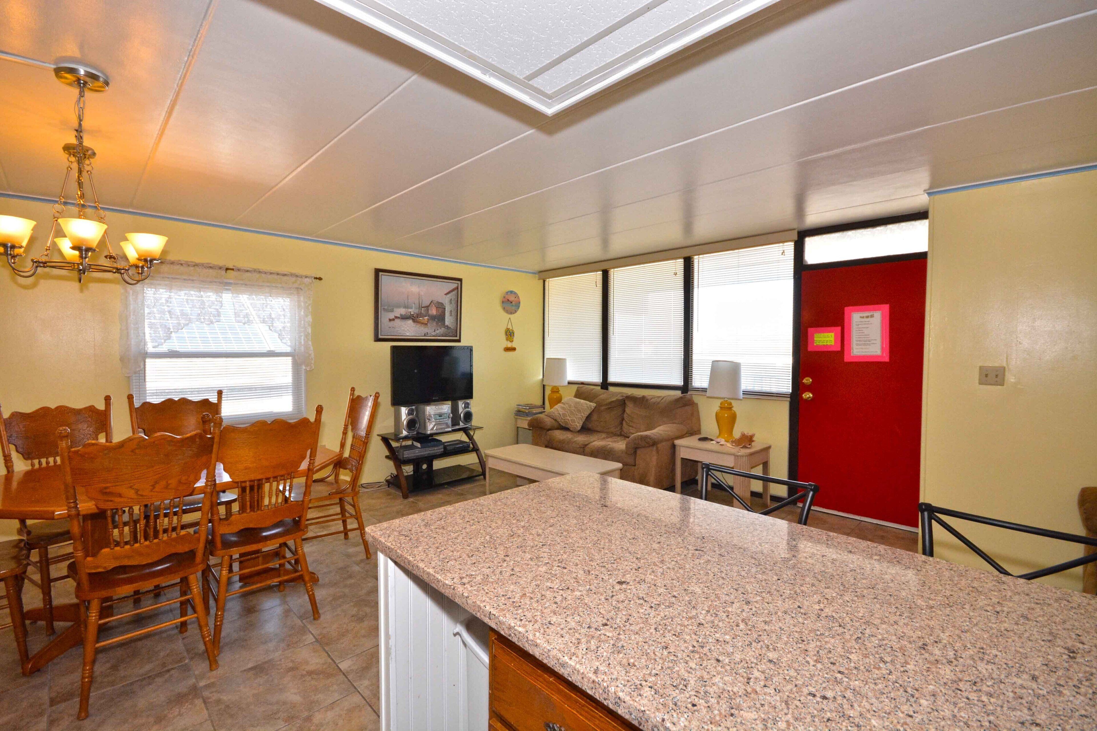 A dining area with wooden chairs and a table, a kitchen with granite countertops, a living room with a couch and TV, and a red door.
