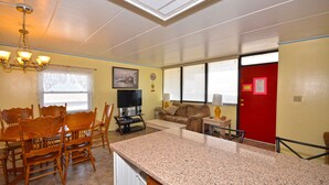 Un comedor con sillas de madera y una mesa, una cocina con encimeras de granito, una sala de estar con sofá y televisión, y una puerta roja.
