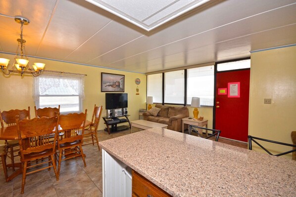 A dining area with wooden chairs and a table, a kitchen with granite countertops, a living room with a couch and TV, and a red door.