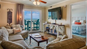 Een woonkamer met een bank, een salontafel en een televisie. Er is een balkon met uitzicht op de oceaan.