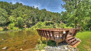 Une terrasse en bois avec des marches menant à la rive d’une rivière, le tout entouré d’une végétation luxuriante et d’arbres.