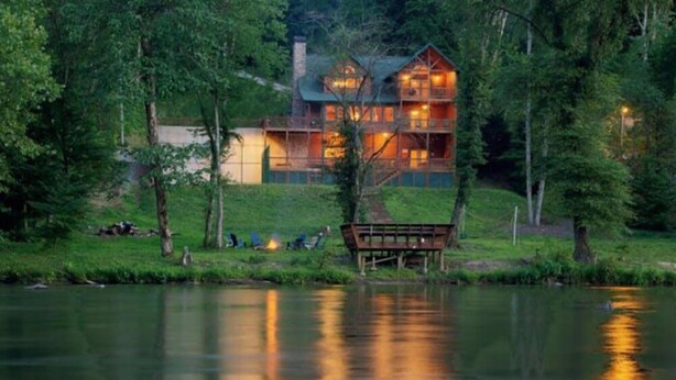 Un chalet au bord du lac, doté d’une grande terrasse et de plusieurs niveaux, entouré d’arbres et bordant un lac paisible.