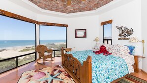 A bedroom with a large window overlooking the beach, a wooden bed, a chair, and a ceiling fan. - Whale of a Tale – Adults-Only Beachfront (Puerto Peñasco)
