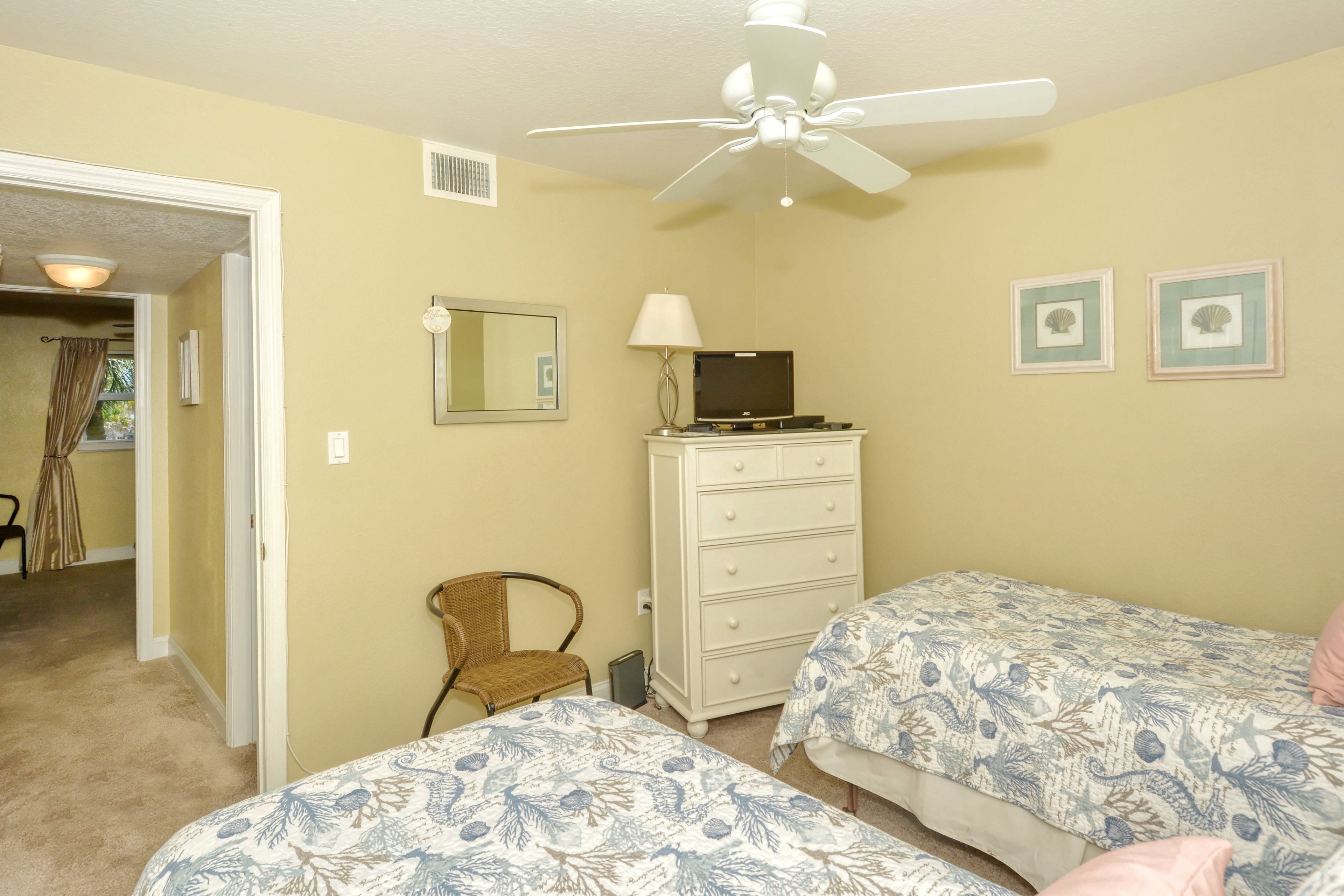 A bedroom with two beds, a dresser, a chair, a ceiling fan, and a door leading to another room.