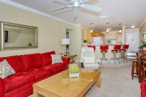 Una sala de estar con un sofá rojo, una mesa de centro de vidrio y un sillón blanco. Hay una zona de cocina con taburetes de bar rojos y una mesa de comedor.