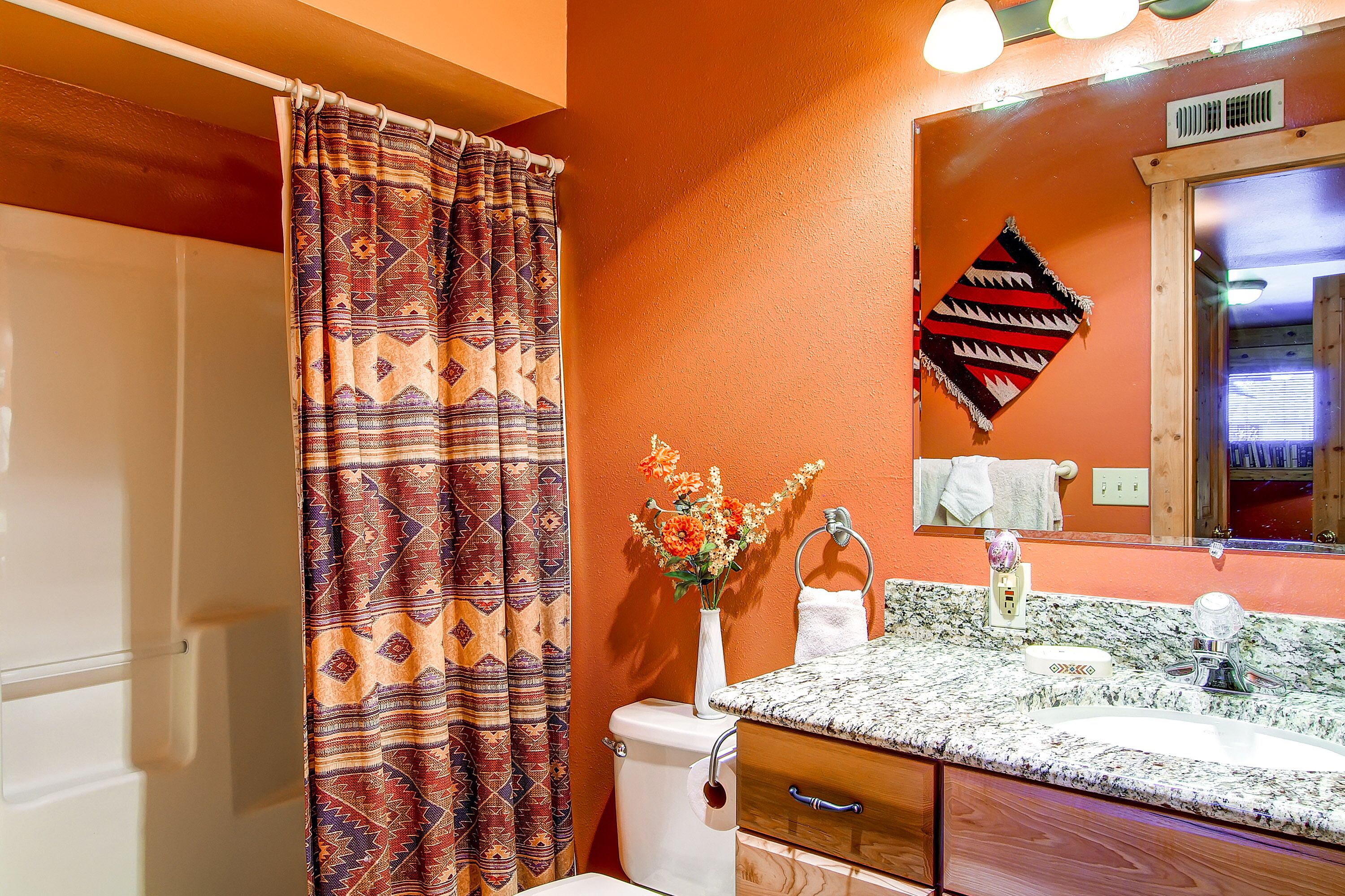 Un baño con cortina de ducha estampada, inodoro, lavamanos con encimera de mármol, un jarrón con flores y un espejo con marco decorativo.