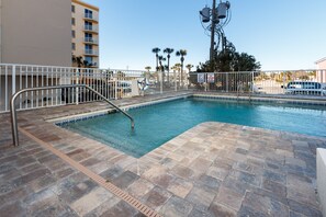 Una piscina con una escalera metálica, rodeada por una cerca blanca, y un edificio con balcones al fondo.