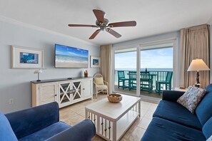 A living room with a ceiling fan, a large TV, a sofa, a coffee table, and a view of the ocean through large windows.