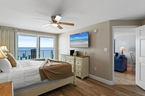 A bedroom with a bed, a ceiling fan, a dresser, a television, and a view of the ocean.