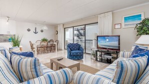Ein Wohnzimmer mit gestreiften Sofas, einem Couchtisch, einem Fernseher, einem Essbereich mit Stühlen und einer durch die Glastür sichtbaren Treppe.
