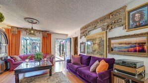 A living room with a purple sofa, a coffee table, and a record player. There are framed pictures on the wall and a large window with curtains. - Bohemian Wonderland: Benicia Bungalow w/ Pergola! (Benicia)