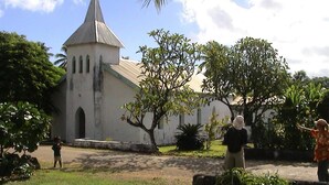 Une église dotée d’un clocher en flèche, entourée de verdure et de palmiers.