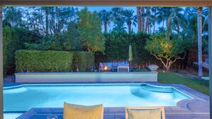 A poolside dining area with a table set for two, a fire pit, and a view of the pool and surrounding greenery. - The Nest – Mid-Century Palm Springs Retreat (Palm Springs)