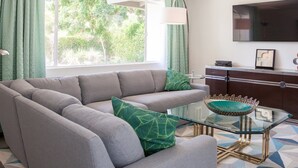 A living room with a sectional sofa, a glass coffee table, a TV, and a view of greenery outside. - The Nest – Mid-Century Palm Springs Retreat (Palm Springs)