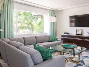 A living room with a sectional sofa, a glass coffee table, a TV, and a view of greenery outside.