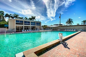 A swimming pool with lounge chairs, a building with multiple floors, and palm trees in the background.
