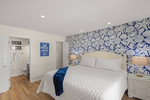 A bedroom with a bed, bedside lamps, a nightstand, a wall with a blue and white pattern, and a door leading to a bathroom.