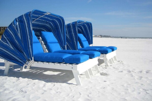 Une rangée de chaises de plage blanches, dotées de coussins bleus et de housses à rayures, est disposée sur une plage de sable.
