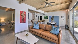 A modern living room with a brown leather sofa, white coffee table, and a ceiling fan. There is a dining area with chairs and a table in the background.