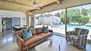 A spacious living area with a brown leather sofa, patterned armchair, and a white coffee table. A large window offers a view of a pool and outdoor seating.
